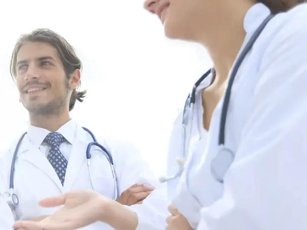 Zwei Gesundheitsfachkräfte, die in die Ferne blicken. Nur ein Gesicht ist sichtbar, das lächelt, während vom anderen nur der Oberkörper zu sehen ist.