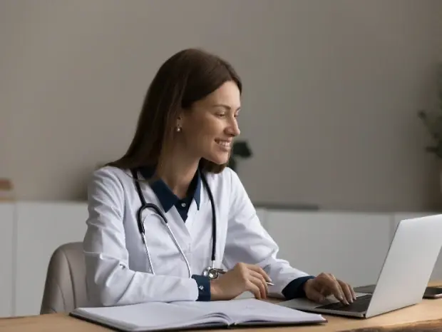 Eine Ärztin, die an einem Tisch sitzt, auf einen Computer blickt und lächelt.