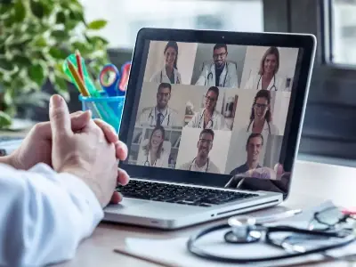 Die Hände einer Person vor einem Laptop, der eine Videokonferenz mit mehreren Teilnehmern zeigt, von denen einige weisse Laborkittel tragen. 