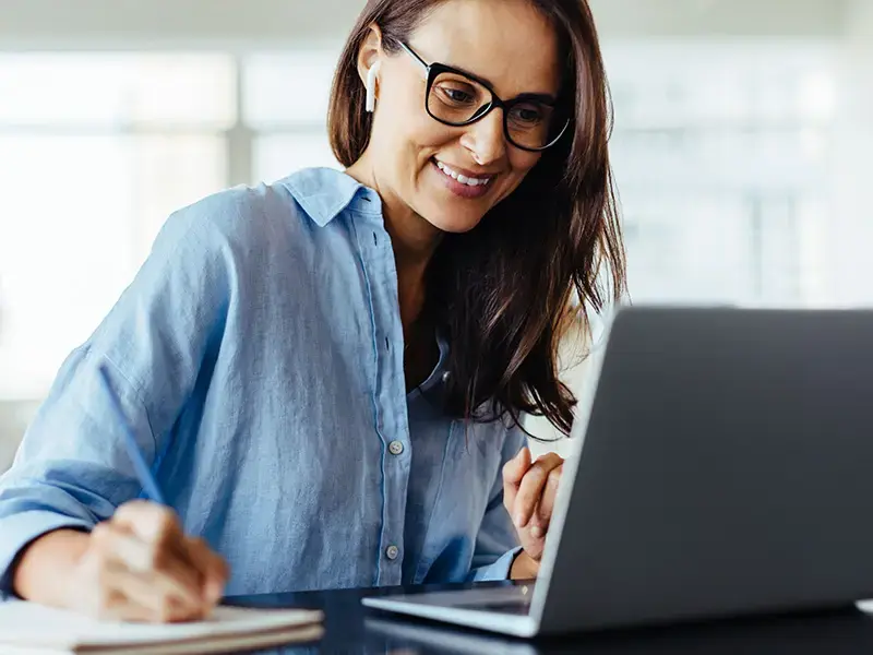 Eine lächelnde Frau mit Brille und hellblauem Hemd, die in einer Büroumgebung an einem Laptop arbeitet.