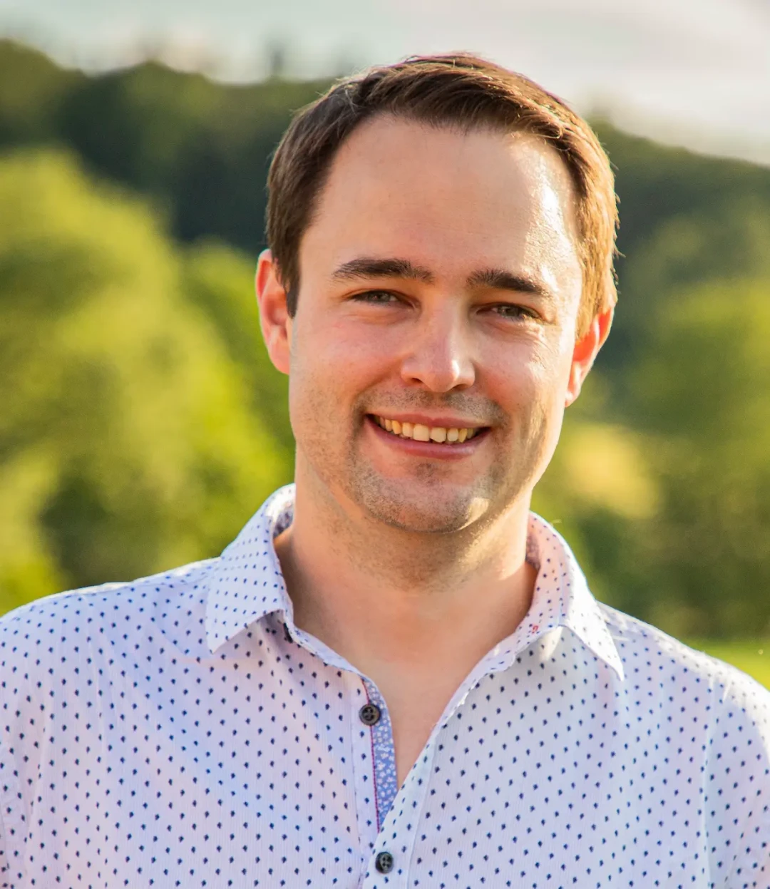 Porträtfoto von Dr. Peter Klar Schubert Florian, Leiter der Primärversorgung Wienerwald in Breitenfurt bei Wien. Er trägt ein weißes Hemd mit kleinen dunklen Punkten und lächelt freundlich. Der Hintergrund zeigt eine unscharfe grüne Landschaft im Freien.