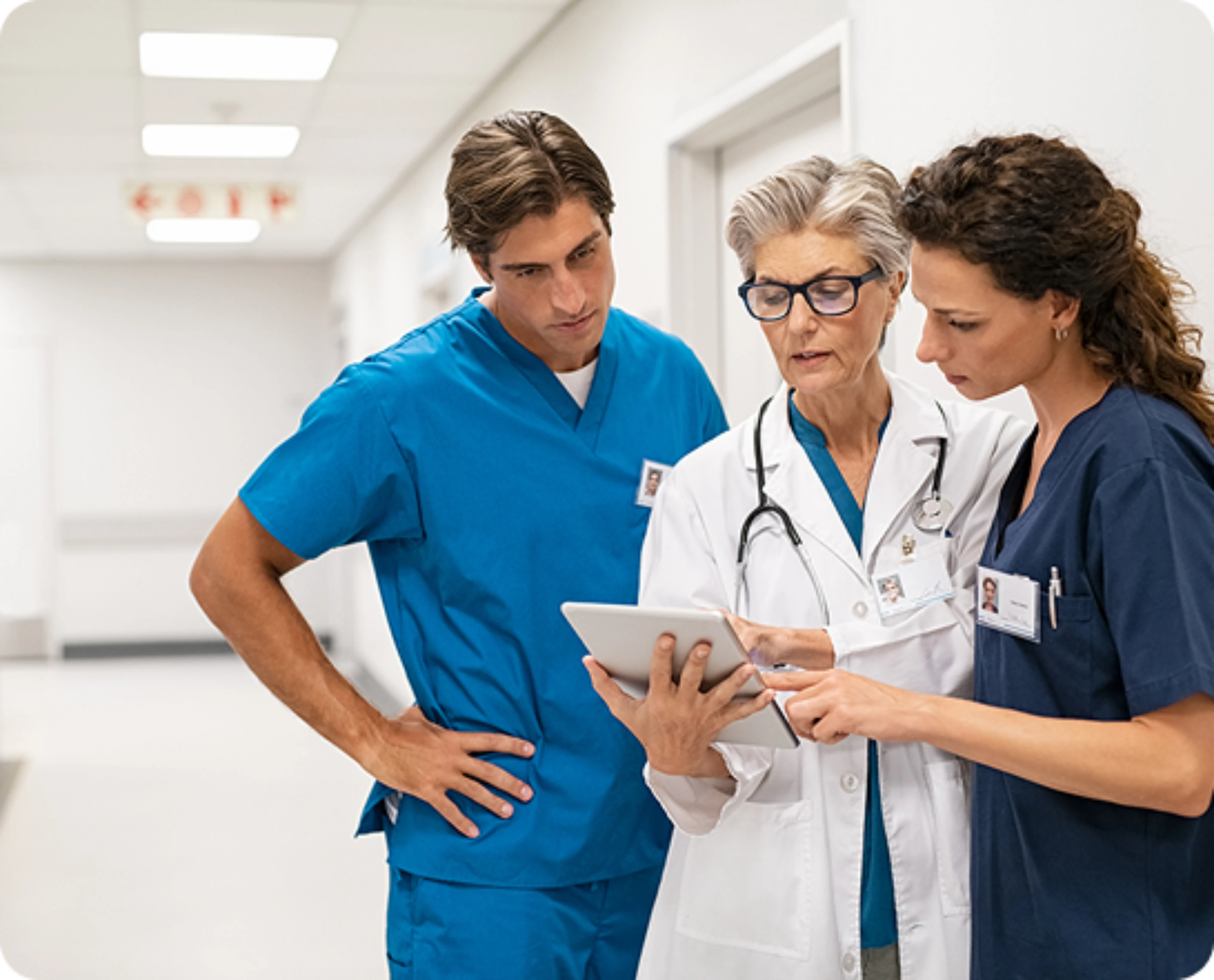 A imagem retrata uma cena num corredor hospitalar com três profissionais de saúde envolvidos numa discussão enquanto observam um tablet.