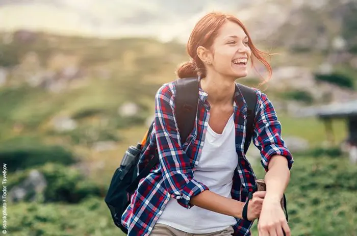Eine lachende Frau trägt einen Rucksack beim Wandern, mit einem bergigen Hintergrund.