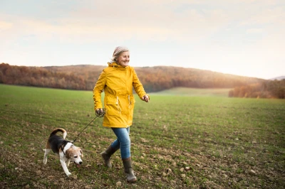 Eine Frau in gelber Regenjacke führt ihren Hund durch eine grüne ländliche Landschaft und geht über ein Feld unter klarem Himmel.