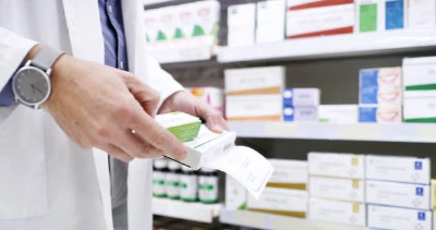 Un pharmacien en blouse blanche tient une boîte de médicaments, avec en arrière-plan des étagères contenant divers produits pharmaceutiques.